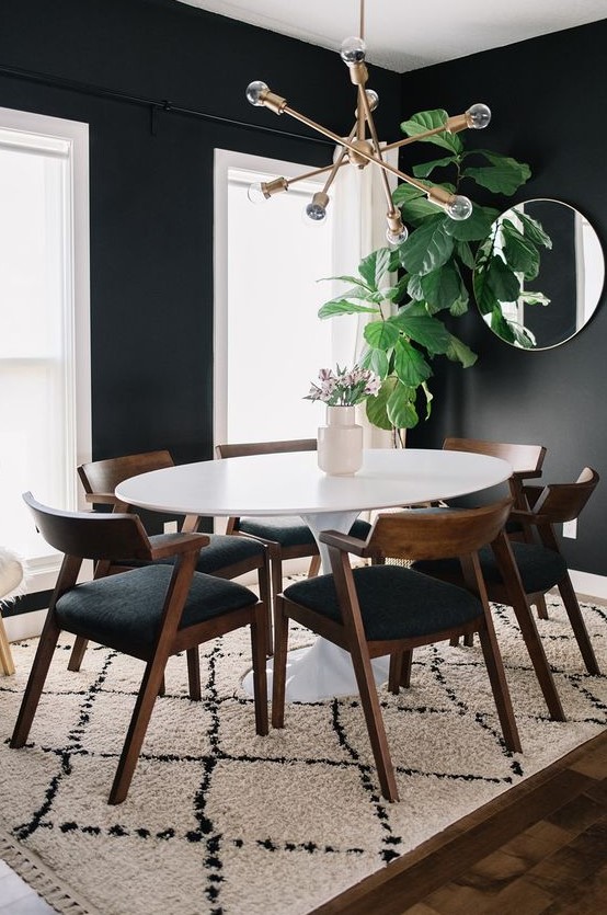 A stylish mid century modern dining room with black walls and white window frames, an oval table and black chairs, a round mirror and a gilded chandelier