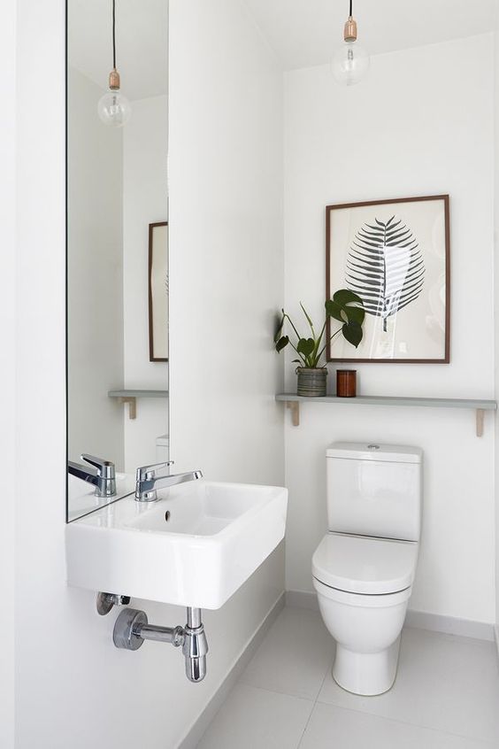 An all white guest toilet with a wall mounted sink, a tall mirror, a plant and a tropical artwork