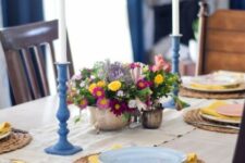 a colorful spring table setting with a bright floral centerpiece, wicker chargers, blue candle holders, yellow napkins