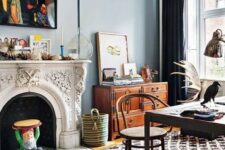 a boho eclectic home office with blue walls, an ornated fireplace, printed layered rugs, a dark desk and a rattan chair, a stained sideboard