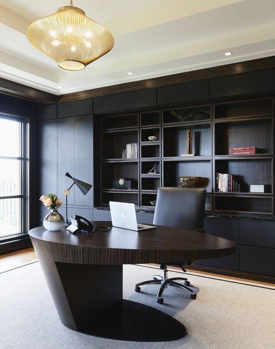 A minimalist and refined home office with a black built in shelving unit, a sculptural oval desk of rich stained wood, a bold chandelier