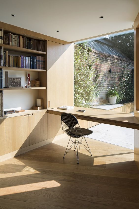 A minimalist home office with a built in plywood storage unit with open storage compartments, a windowsill desk and a black chair