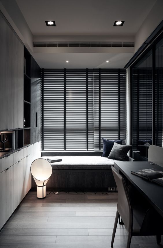 A moody contemporary home office with a windowsill daybed, a built in storage unit, a black desk and black chairs and some lights