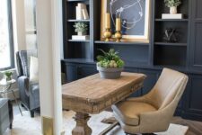 a pretty farmhouse home office with a grey storage unit, a stained rustic desk, a tan leather chair and artworks, a gilded chandelier