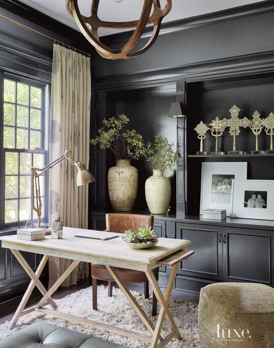 A stylish home office with black walls, black storage units, a light stained desk and a brown chair, a wooden sphere chandelier
