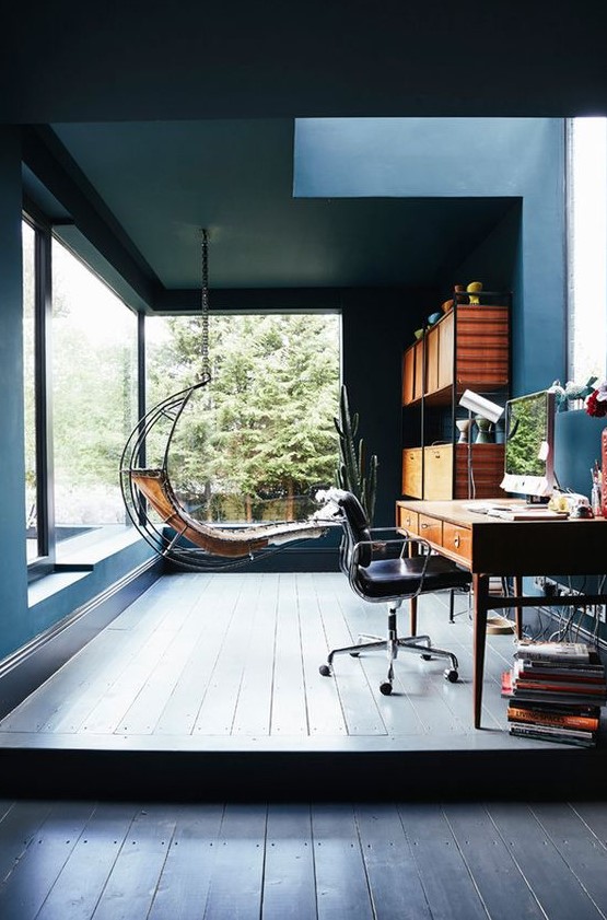a welcoming home office in blue, with panoramic windows and a skylight, a wooden desk plus a leather chair, a hanging lounger and a shelving unit