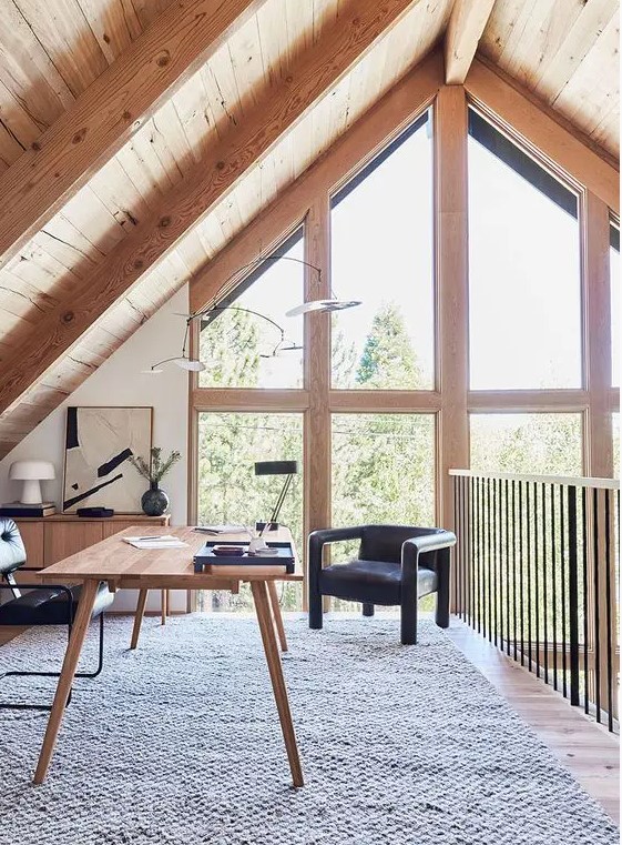 An attic home office with a wooden desk and a storage unit, black chairs, some lamps and a gorgeous view through double height windows