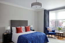 a stylish bedroom with light grey walls brought to life with medium blue curtains, armchair and throw, contrasted with burnt orange cushions on the bed