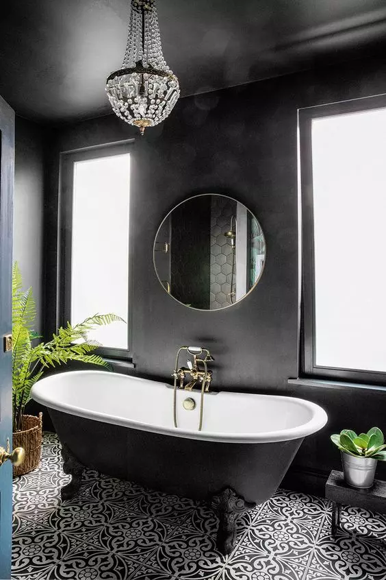 a black bathroom with a black and white mosaic tile floor, a black clawfoot tub, a crystal chandelier and potted greenery
