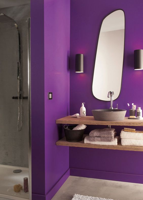 a bright contemporary bathroom with a purple part and a grey shower space, a floatign wooden vanity and a catchy-shaped mirror