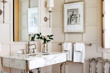 a chic beige bathroom done with wall panels, elegant furniture and lamps and a marble sink on tall legs