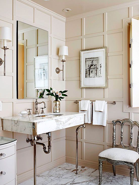 a chic beige bathroom done with wall panels, elegant furniture and lamps and a marble sink on tall legs