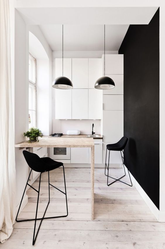 a minimalist monochromatic kitchen with white sleek cabinets, blakc pendant lamps, a plywood countertop, black stools and a black statement wall