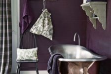 a moody rustic bathroom done in deep purple, with a metal tub, vintage shelves and a chair plus plaid curtains
