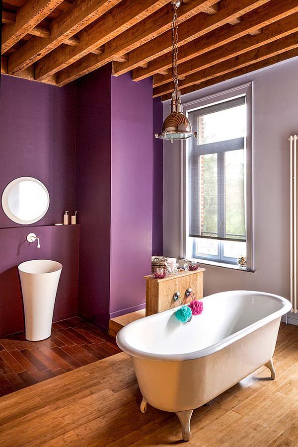 A purple farmhouse bathroom with a wooden ceiling with beams, a pendant lamp, a free standing sink and a vintage bathtub