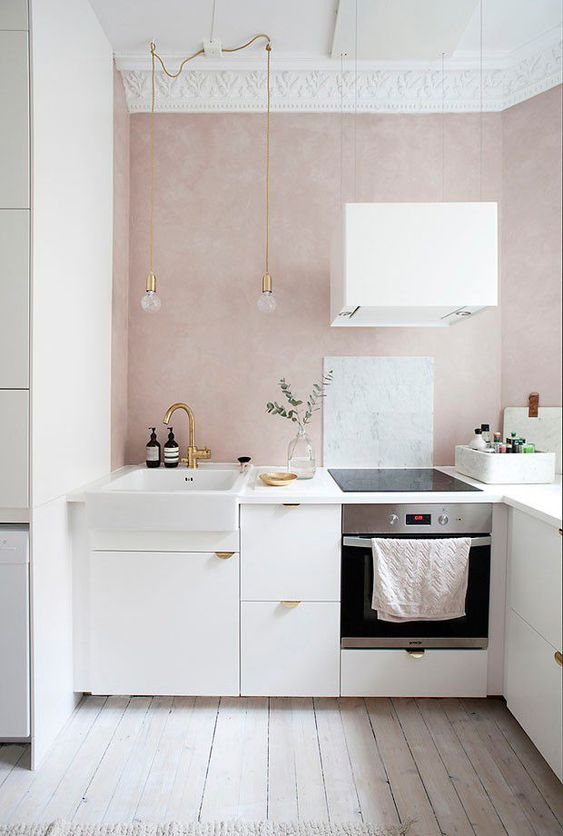 a small and serene contemporary kitchen with blush walls, white cabinets with gold pulls, pendant bulbs and a white hood