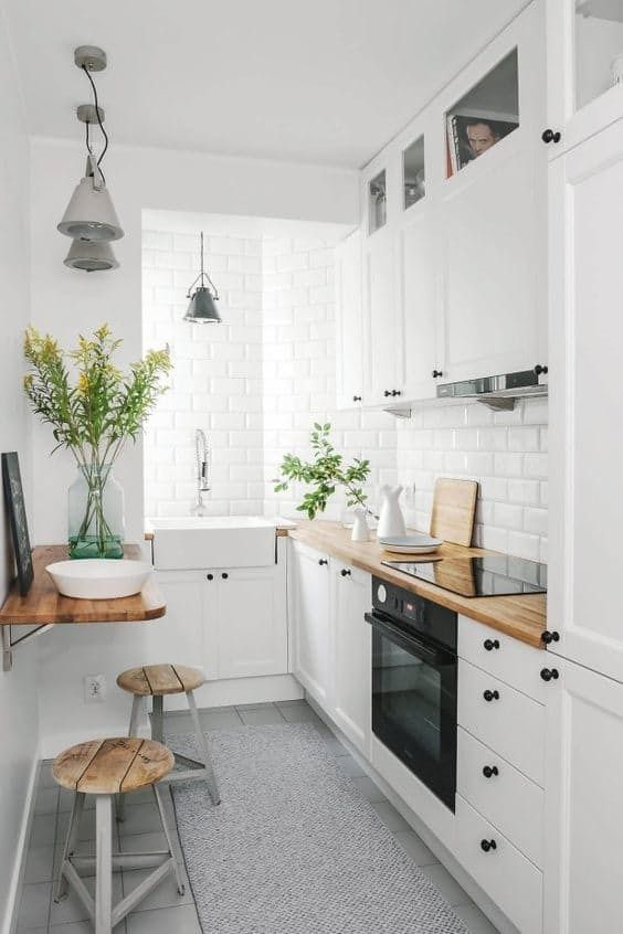 a small contemporary kitchen in white with walls done with tiles, a folding mini table, metal and wood stools and pendant lamps