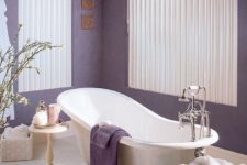 a stylish bathroom with purple walls and towels, an elegant vintage bathtub, side tables and a wicker box