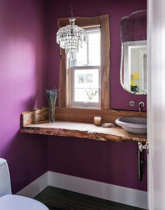 an eclectic bathroom with purple walls, a wooden vanity with a livign edge, a crystal chandelier and a lavender vessel sink