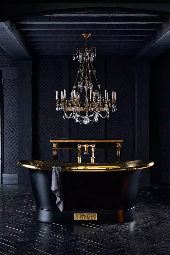 an exquisite black and gold bathroom clad with tiles and done with textural wallpaper, with a black tub and a chic chandelier with crystals