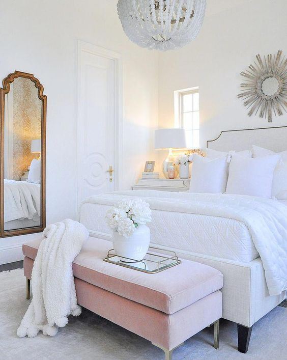 a chic glam bedroom in white with an upholstered bed and bench, a vintage mirror, a crystal chandelier and a sunburst mirror