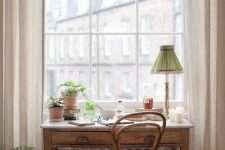 a cute working nook by the window with a stained desk and a chair, a table lamp and some greenery in pots plus a view
