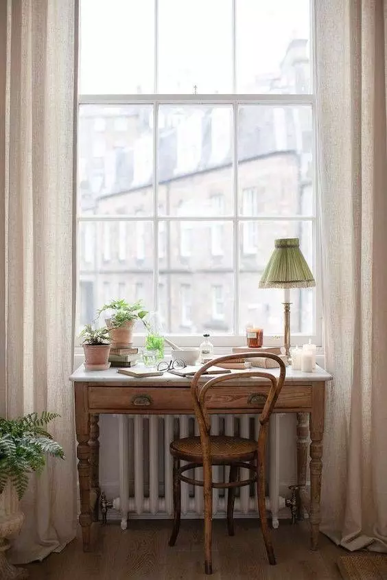 a cute working nook by the window with a stained desk and a chair, a table lamp and some greenery in pots plus a view