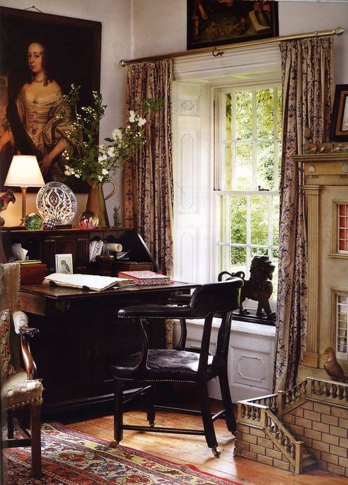 a fab vintage home office with vintage artwork, a dark-stained bureau desk and a black chair, printed curtains and a rug plus some blooms