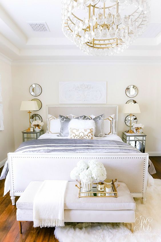 a luxurious glam bedroom in neutrals with a crystal chandelier, mirror nightstands, an upholstered bed, mirrors and a tray