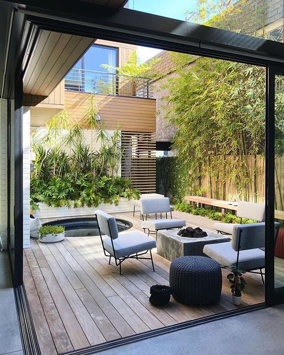 a modern patio with lots of greenery, a hot tub, a concrete firepit, grey chairs and a wicker ottoman