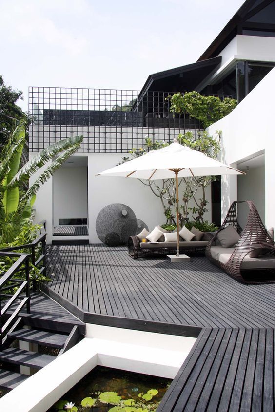 a modern terrace with a dark wooden deck, dark wicker furniture, neutral pillows and lots of planter greenery and trees around