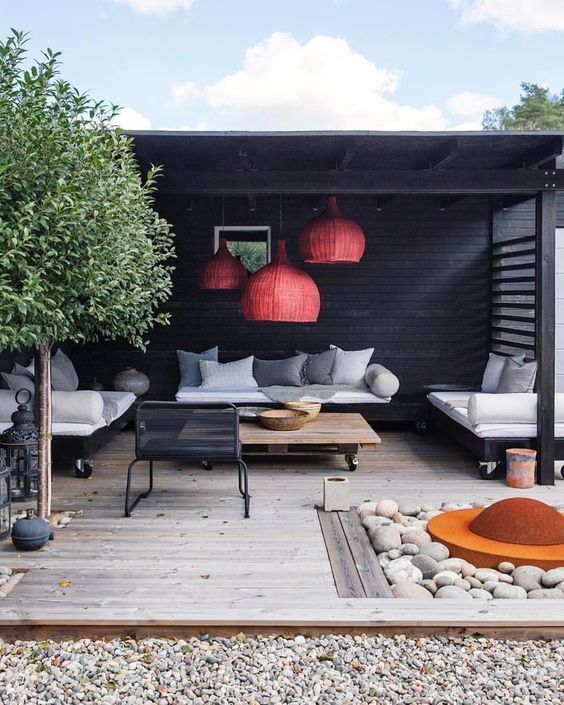 a modern terrace with red wicker lamps, modern furniture on casters, a fire pit with pebbles around and a tree