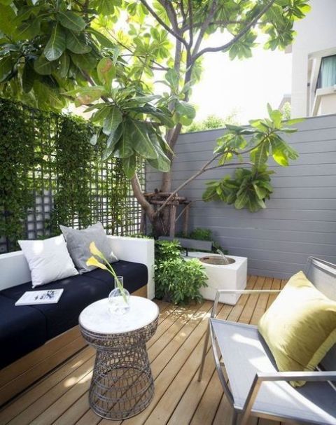 a modern terrace wityh wood and metal furniture, a wire table, greenery and a tree with a fountain