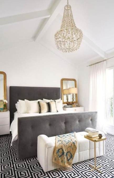 a monochromatic bedroom with gold touches, a crystal chandelier, gold frame mirrors and a table, a grey upholstered bed
