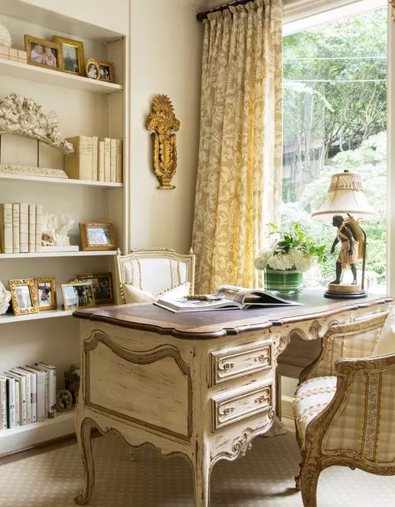 a neutral home office with built-in bookshelves, a refined shabby chic desk, matching refined chairs and a table lamp plus floral curtains