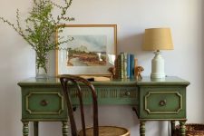 a small and chic vintage working space with a green desk, a stained chair, a table lamp, some books and greenery in a vase