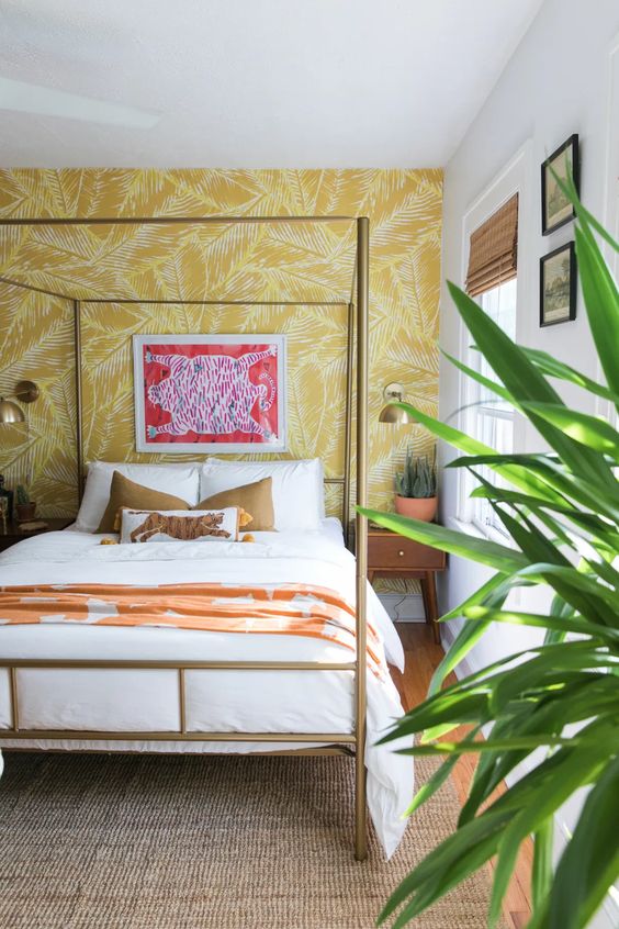 a tropical glam bedroom with a yellow printed wall, a gold bed, potted greenery, colorful pillows and textiles