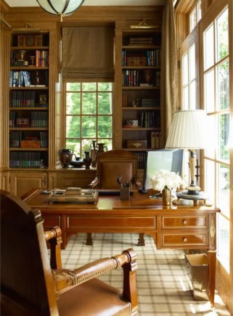 A vintage home office with built in bookcases, a dark stained vintage desk, matching leather chairs, a table lamp and some blooms
