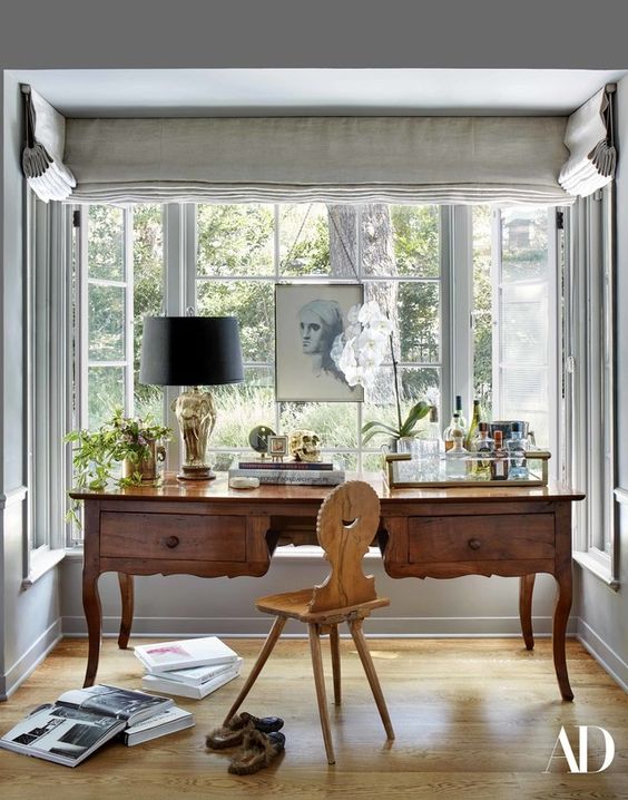 A vintage working nook by the window, with a large stained desk and a light stained chair, a vintage table lamp and some art