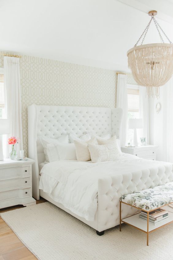 a white glam bedroom with a statement upholstered bed, white and floral furniture, a fringe chandelier