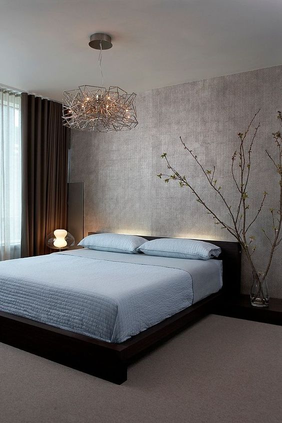 A zen like bedroom with a silver accent wall, a dark bed with blue bedding, blooming branches and very eye catchy lamps