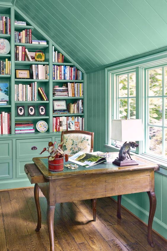 An attic home office with green walls and a ceiling, a built in bookcase and a stained desk, a floral chair and a table lamp
