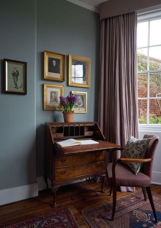An exquisite vintage working nook with grey walls, a vintage dark stained bureau and a mauve chair, mauve curtains and a gallery wall