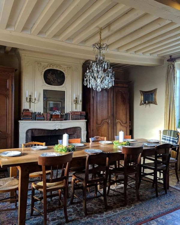 A French country dining space with a non working fireplace, a long stained table and mismatching chairs, a crystal chandelier