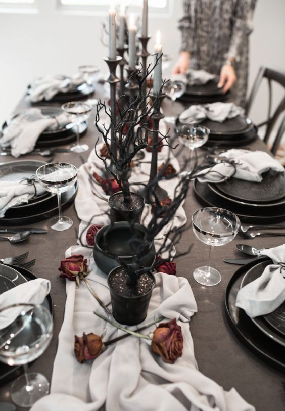 a Halloween tablescape with a white runner, dried roses, black branches, grye candles and elegant black plates