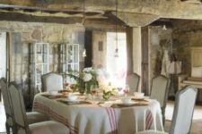 a Provence dining room with a stained ceiling with beams, stone walls, a table and vintage chairs, pendant lamps