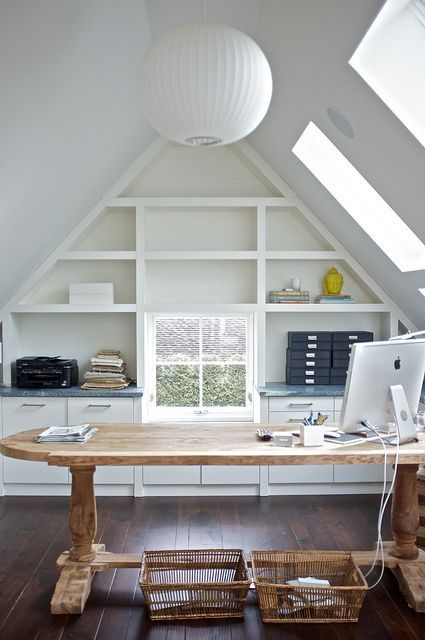 A beautiful attic home office with skylights, a built in shelving unit, a large stained desk, baskets and a sphere pendant lamp