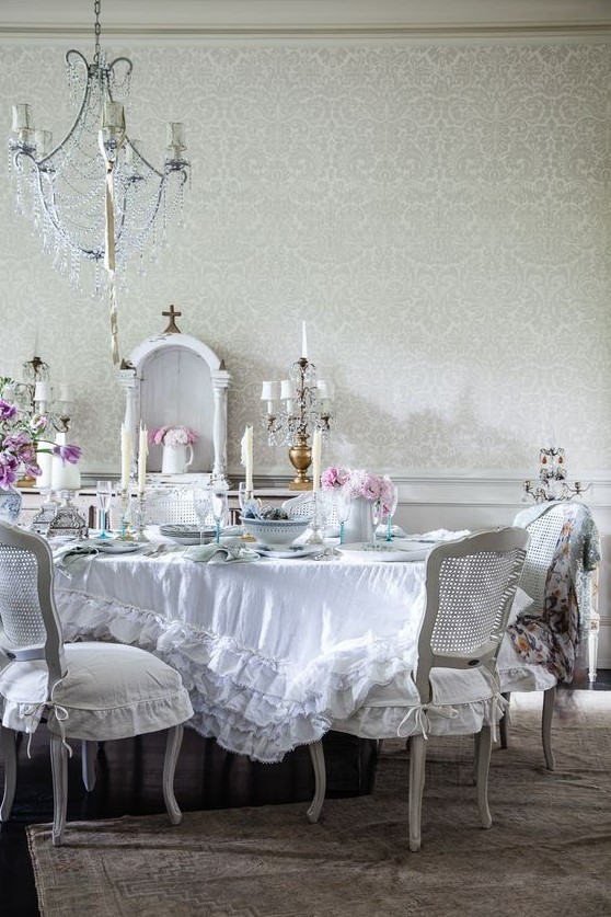 a beautiful shabby chic dining room with a crystal chandelier, white furniture, a ruffle tablecloth and blooms