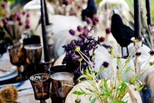a bright and chic Halloween tablescape with white pumpkins, purple blooms, greenery, black candles, crows, dried grasses and goblets