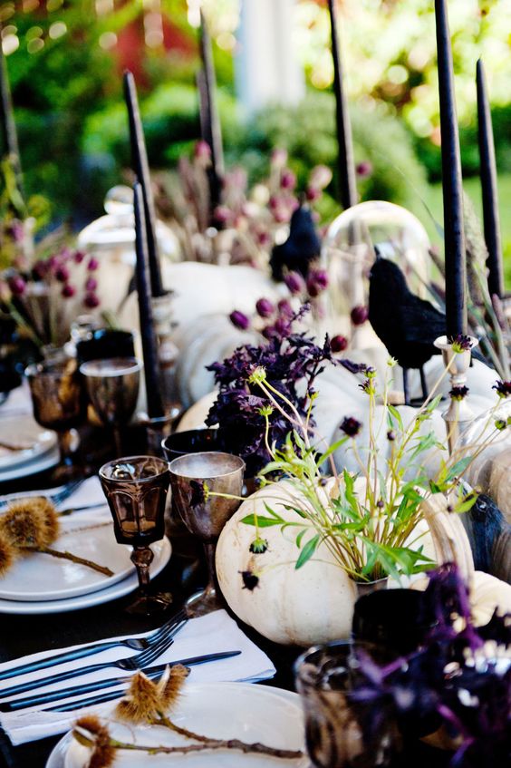 a bright and chic Halloween tablescape with white pumpkins, purple blooms, greenery, black candles, crows, dried grasses and goblets